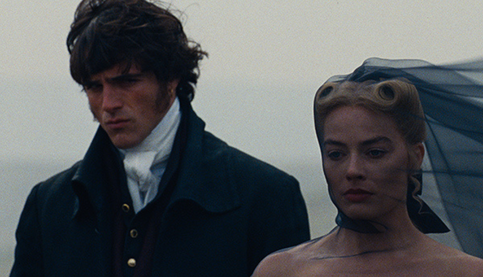 Two people in historical-style clothing stand side by side outdoors on a grey, overcast day. The person on the left wears a dark coat over a high-collared white shirt, while the person on the right wears a dress with a sheer dark veil flowing back from their head.